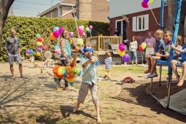 Little girl hitting pinata at birthday party in backyard. Lots of colorful ballons in the background with family and guests. Some kids are sitting on the trampoline watching with interest.