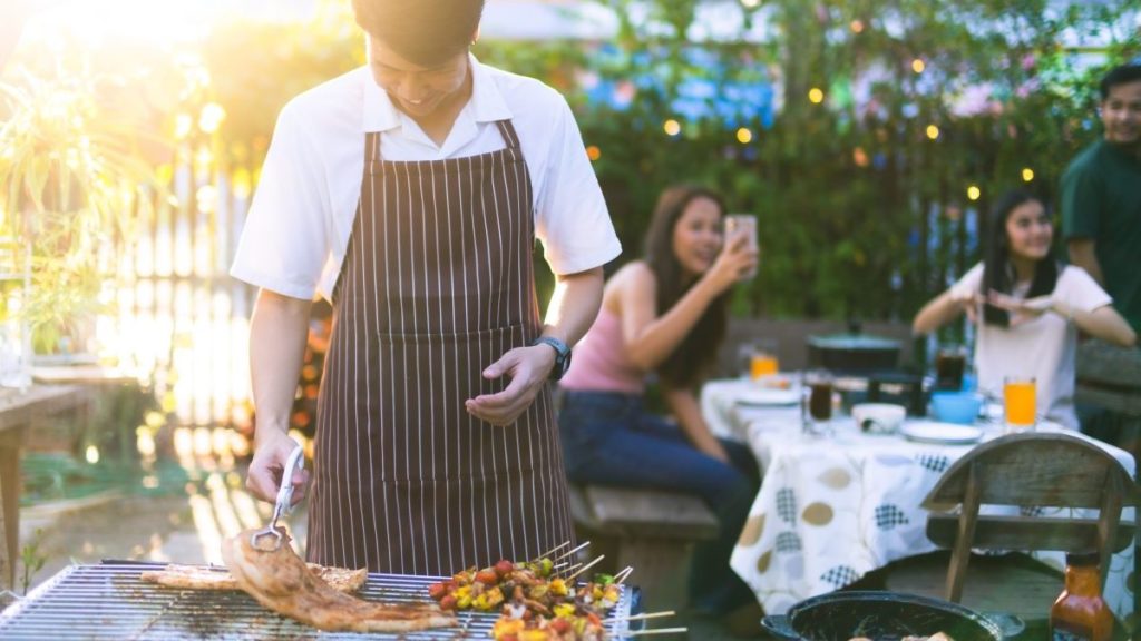 A man happily grilling food as people enjoy the party in the background.