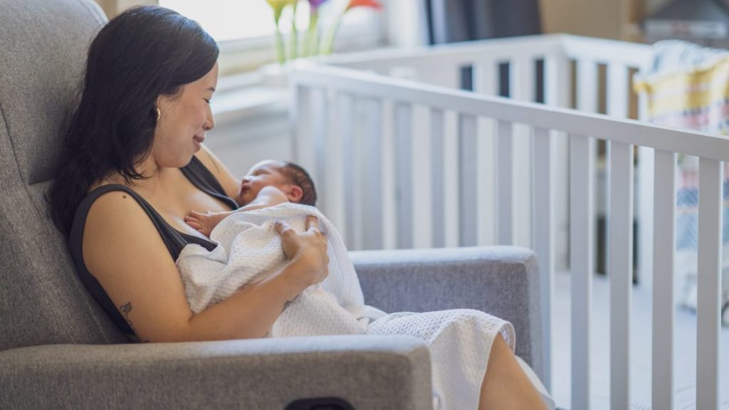 A new mother sits in a rocking chair in her daughter's nursery as she gently rocks her new baby to sleep. She has the baby lightly wrapped in a blanket for warmth as she gazes down at the infant with a smile.