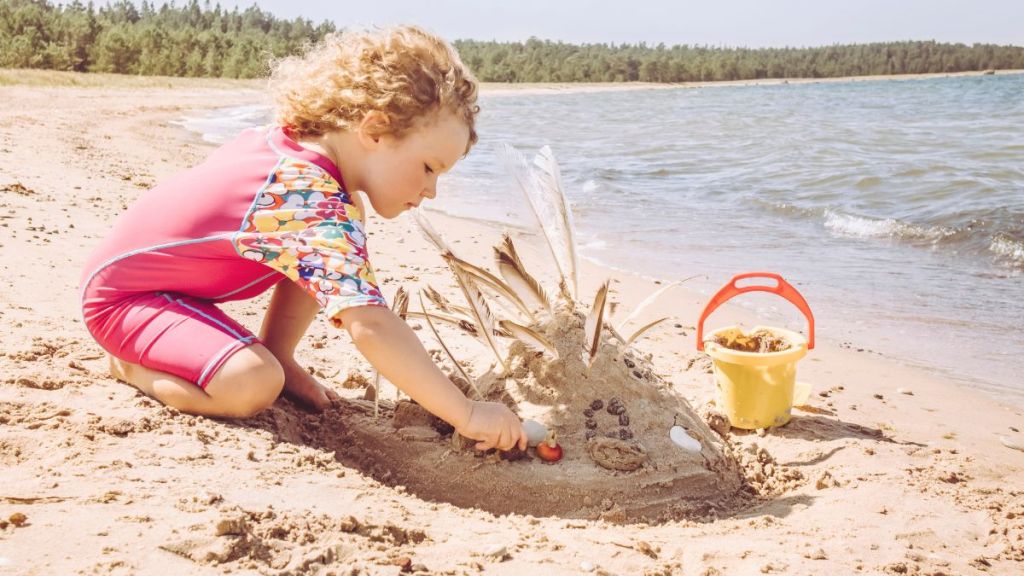 5-year-old girl enjoy play in sand wearing pink long UV protective swimsuit, protecting light sensitive skin from sunburn.