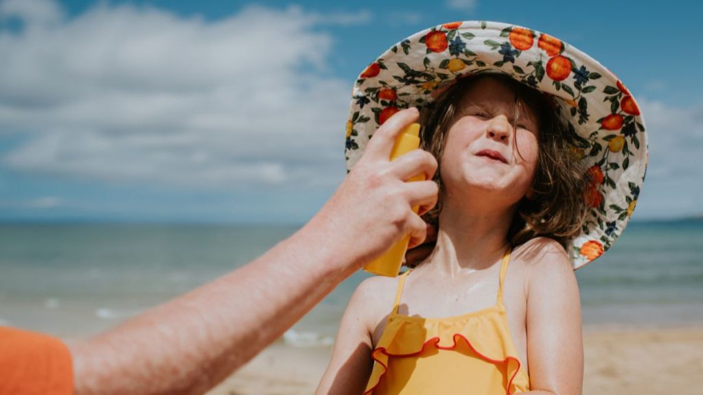 Comical image of a little girl having sunscreen applied to her face. She shuts her eyes tightly and closes her mouth, in anticipation of being spritzed in the face with liquid spray suncream.