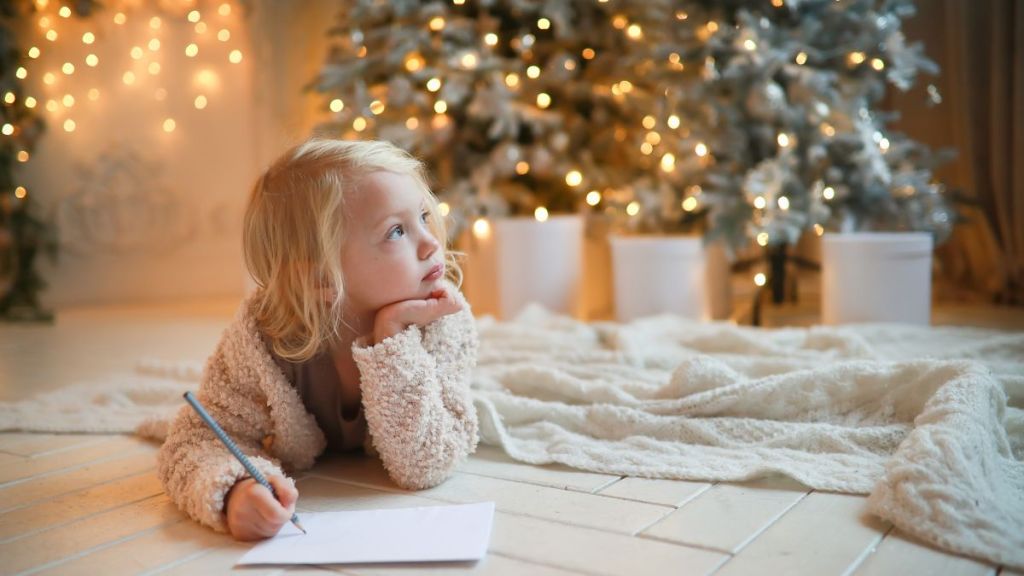 Cute girl child blonde in cozy cardigan writes letter to Santa Claus, a perfect activity for parents and kids, near Christmas tree lying on floor.